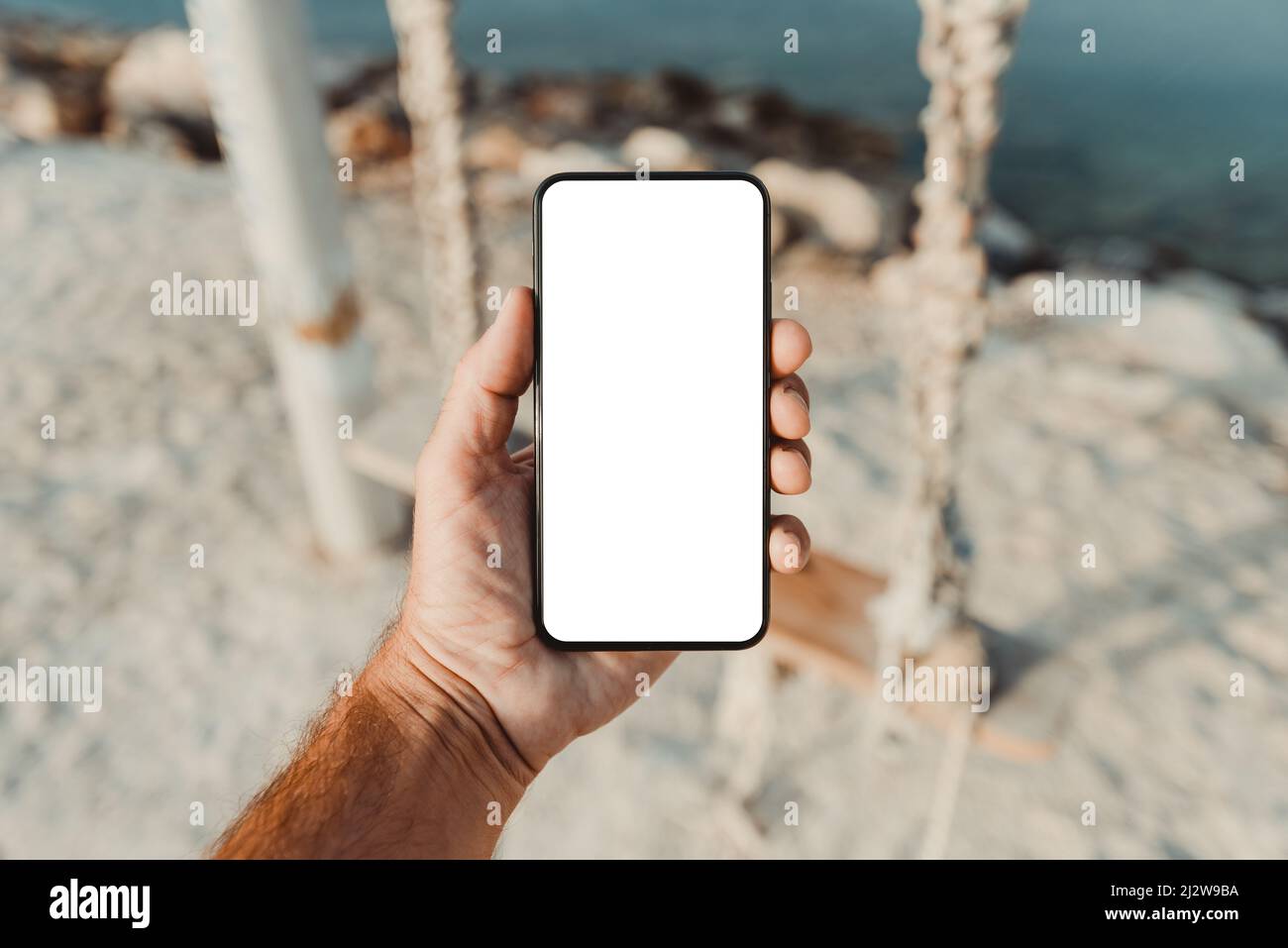 Man standing by the sea and holding mobile smart phone with blank mockup screen, closeup of hand and device, selective focus Stock Photo