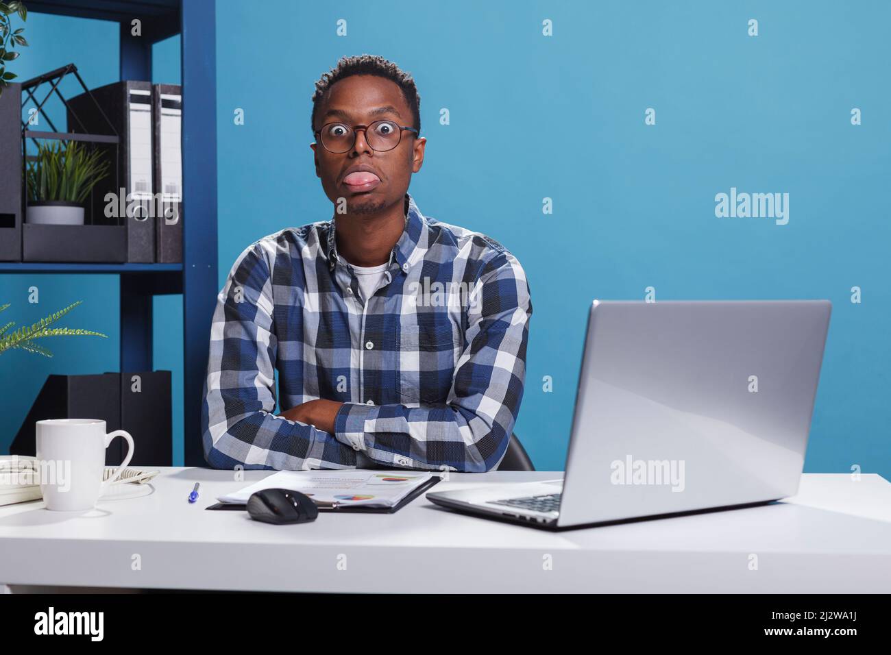 Funny and goofy office working person making amusing playful face while sticking out tongue. Childish and dumb young adult man with dullard face expression while in company workspace. Stock Photo