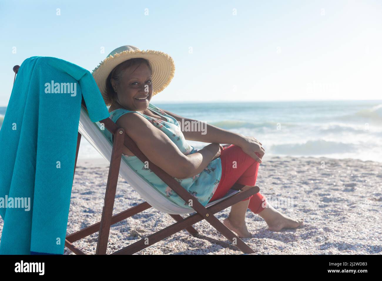 Portrait of smiling african american senior woman wearing hat sitting on folding chair at beach Stock Photo