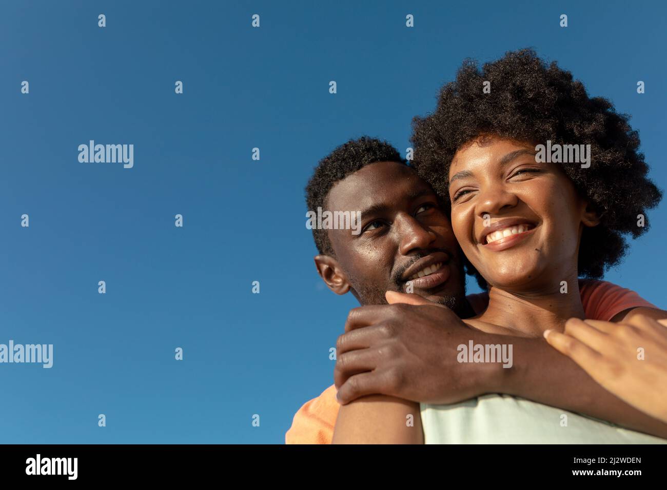 Low angle view of happy african american man hugging girlfriend from behind against clear blue sky Stock Photo