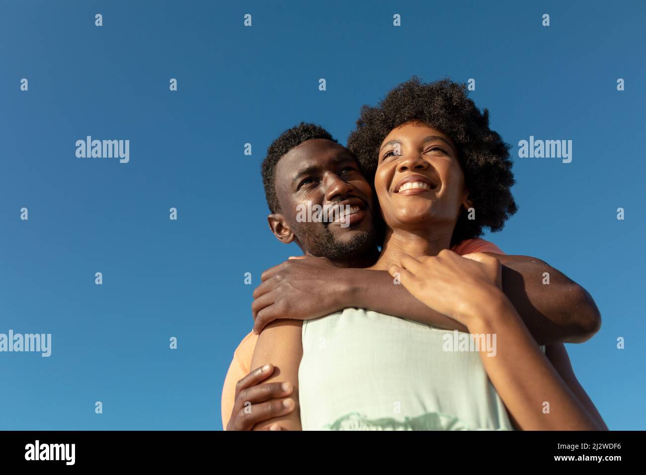 Low angle view of smiling african american man hugging girlfriend from behind against blue sky Stock Photo