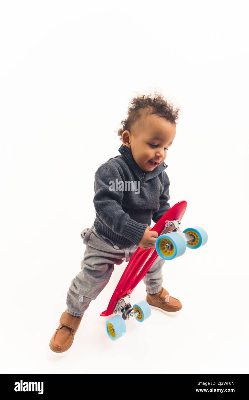 lovely African American little boy standing and holding a pink skateboard wearing brown shoes grey trousers and blue pullover studio shot white background full shot copy space. High quality photo Stock Photo