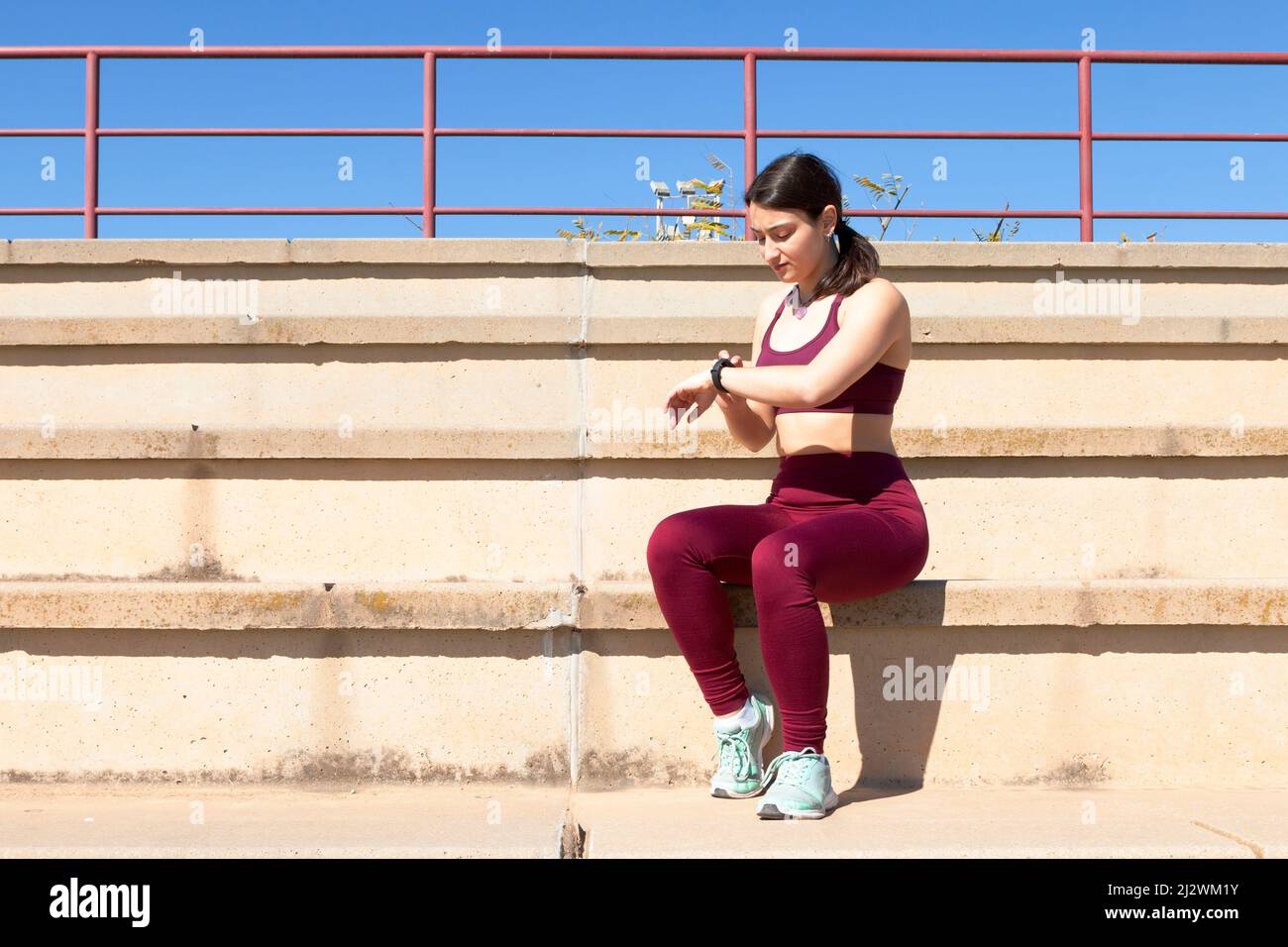 General shot of a young Caucasian female athlete dressed in maroon leggings, a purple top and turquoise sneakers is sitting on a bleacher while touchi Stock Photo