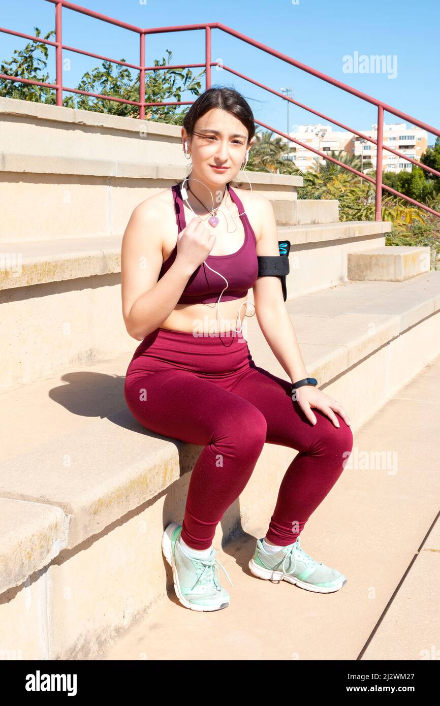 A young Caucasian female athlete dressed in maroon leggings, a purple top and turquoise sneakers is sitting on a bleacher while holding the cord of he Stock Photo