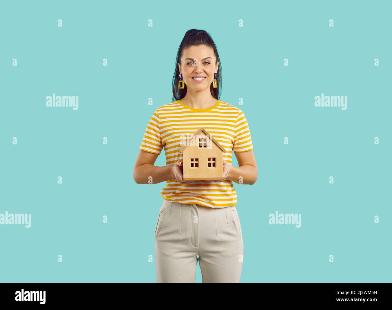 Portrait of pretty smiling woman holding wooden model house standing on blue background. Property investment and house mortgage financial concept. Stock Photo