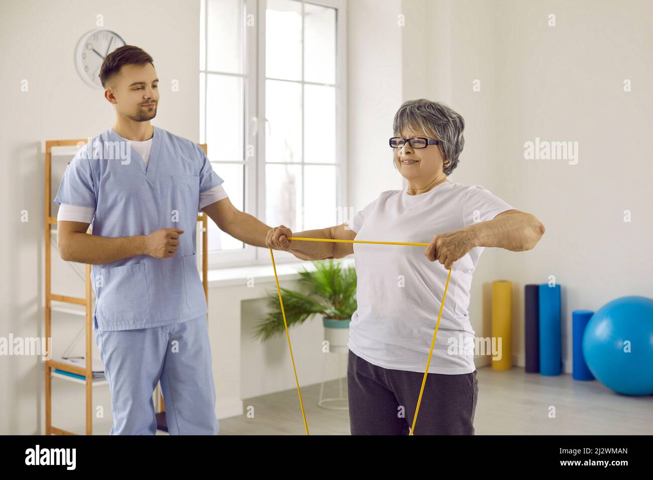 Joyful old woman with doctor trains during rehabilitation with help of resistant band. Stock Photo