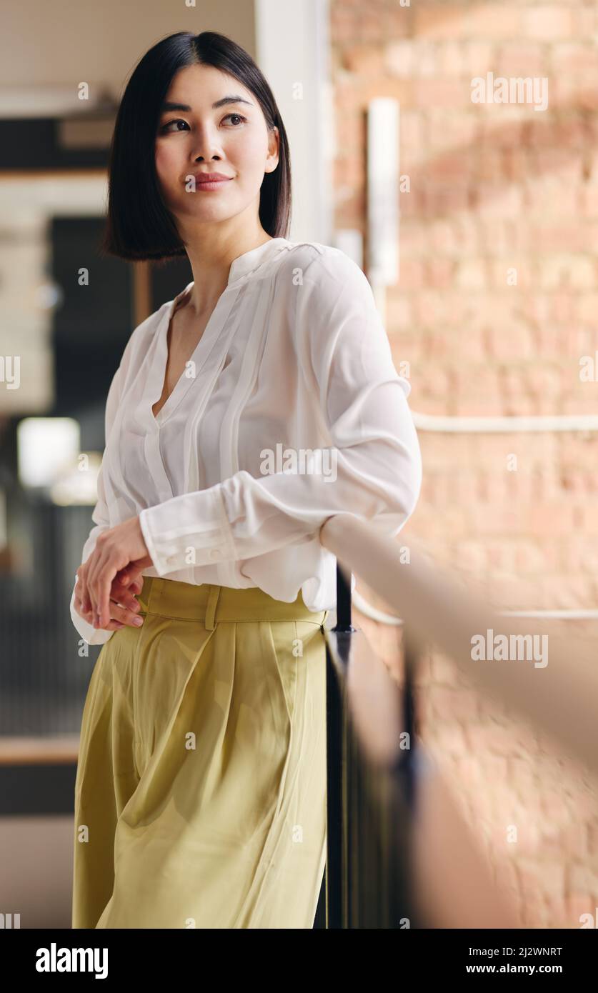 Portrait of confident mid adult Chinese businesswoman leaning on railing and looking away with a serene expression Stock Photo