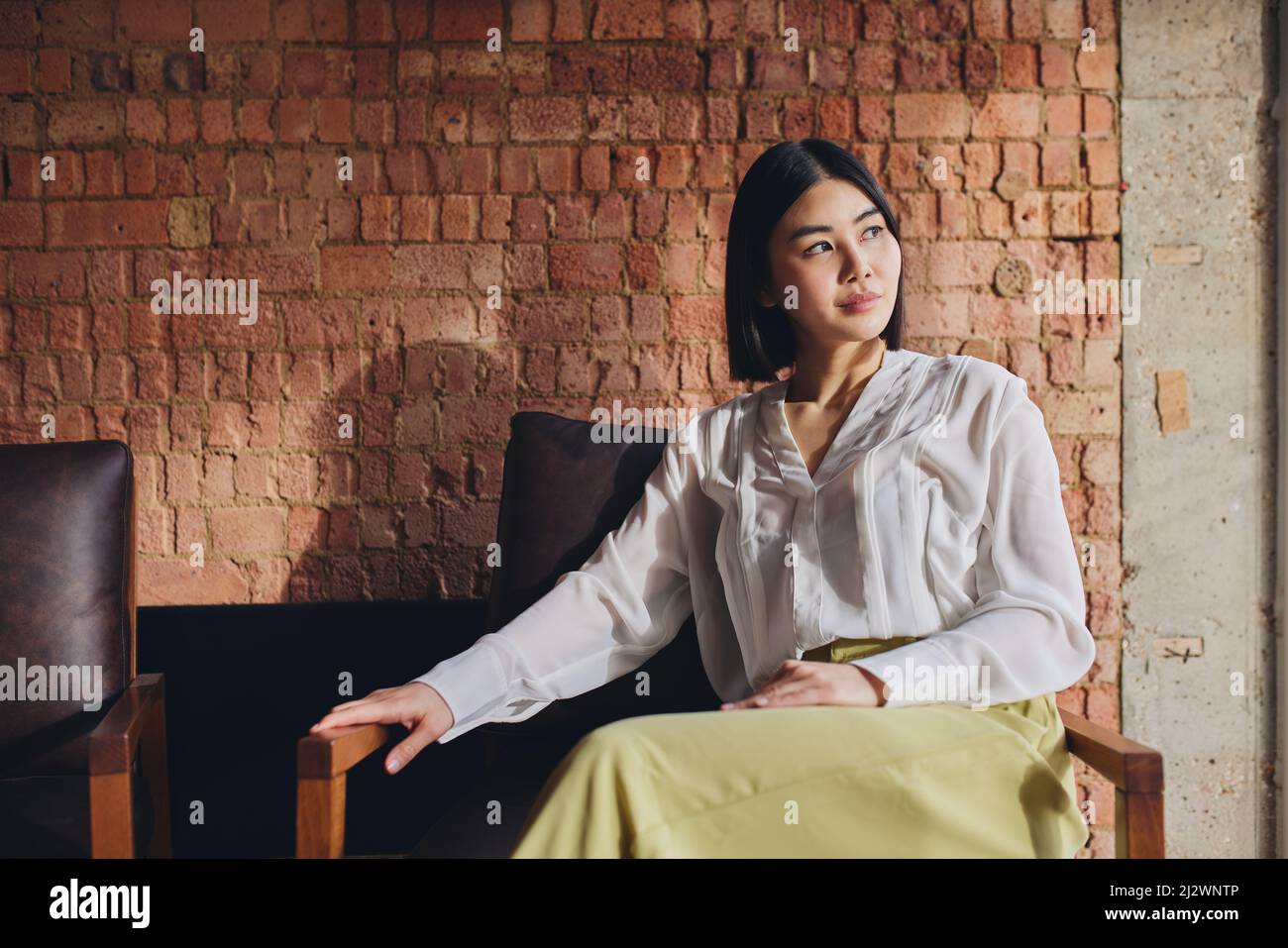 Portrait of mid adult Chinese businesswoman sitting on chair in front of exposed brick wall and looking away Stock Photo