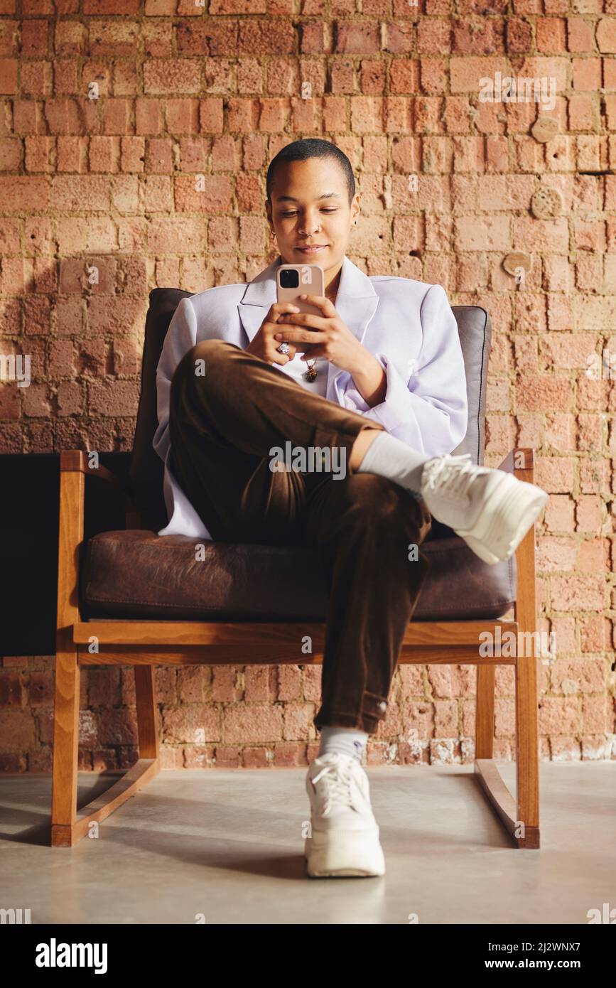 Portrait of confident multiracial LGBTQ mid adult woman sitting on retro chair and using smartphone in front of exposed brick wall Stock Photo