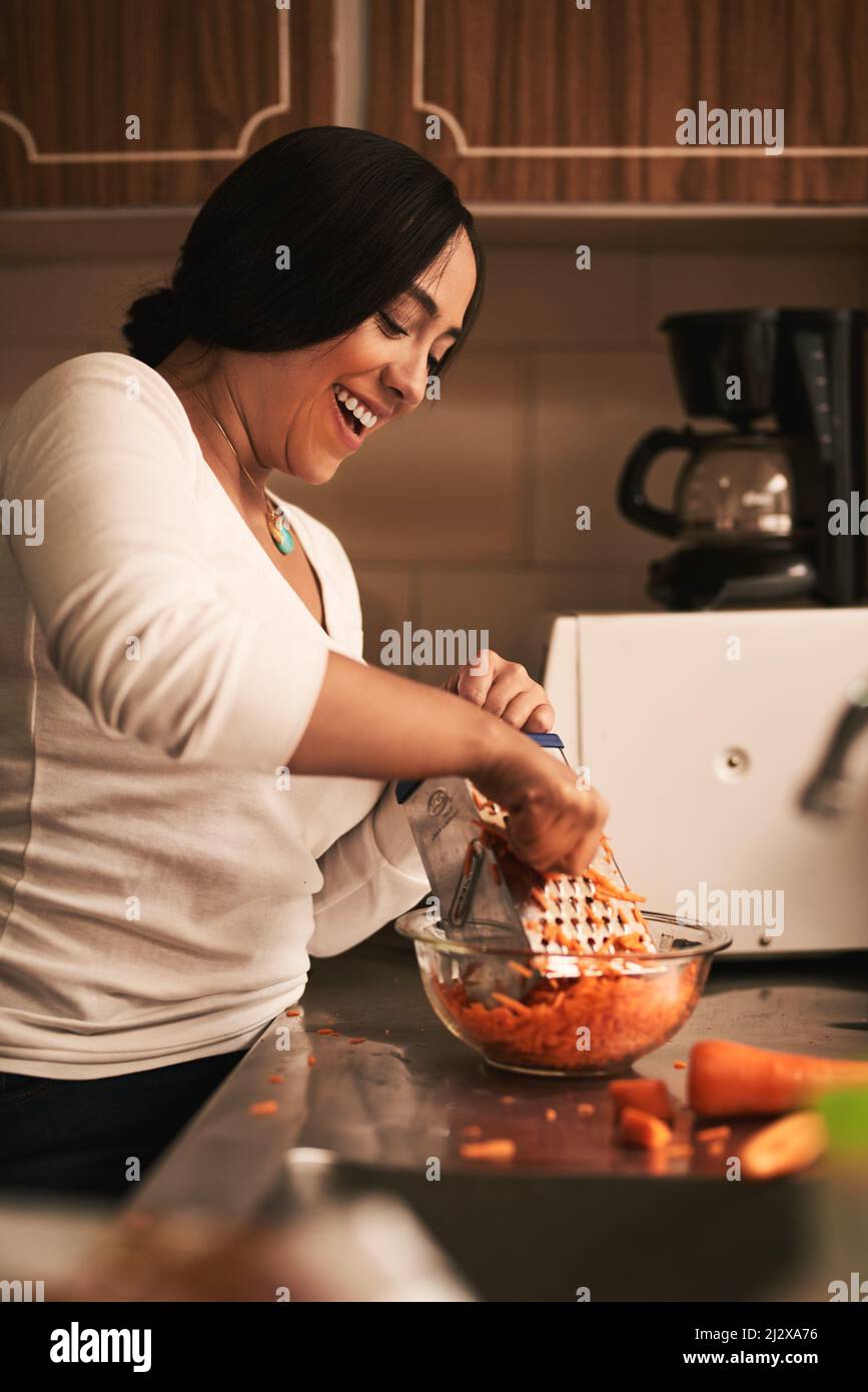 These are going to turn out great. Shot of a cheerful young woman baking cupcakes and grating carrots inside of a kitchen at home. Stock Photo