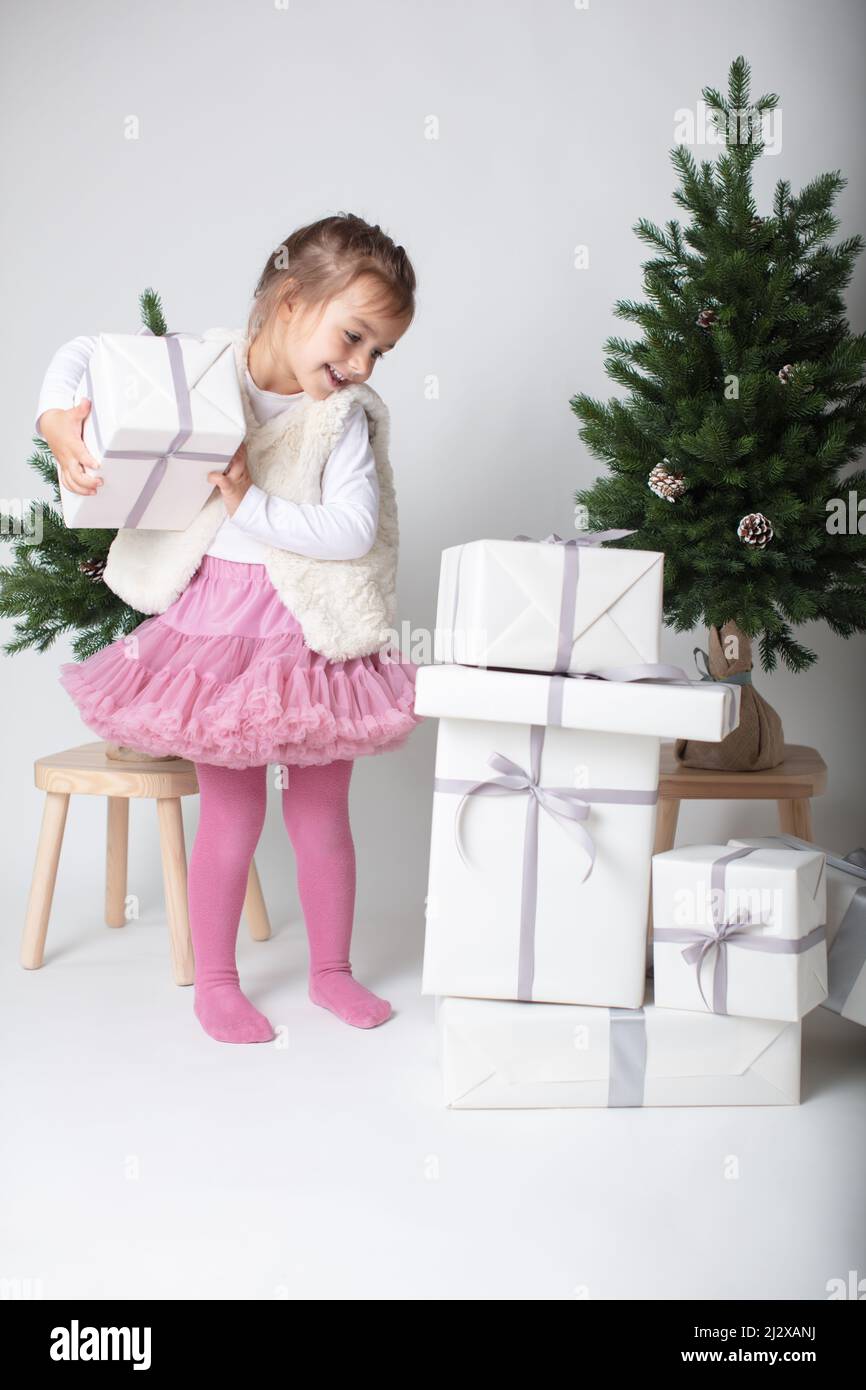 Happy toddler girl selecting her Xmas gift from a pile of gift boxes under Christmas tree. Modern Xmas decoration. High-key image on white background Stock Photo