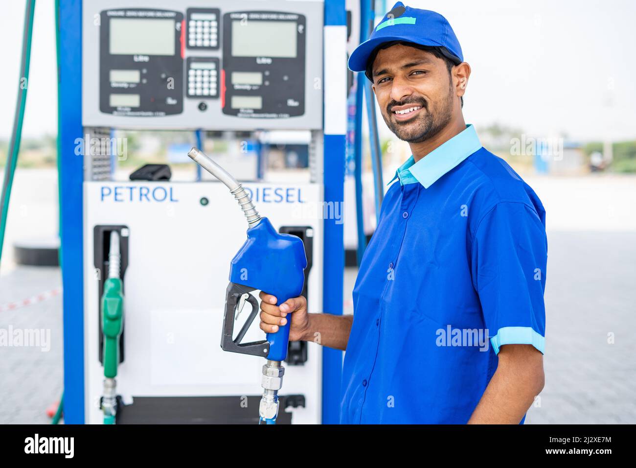 Happy petrol pump worker standing by holding fuel nozzle while looking camera at gas filling station - concept of happiness, job and petroleum service Stock Photo