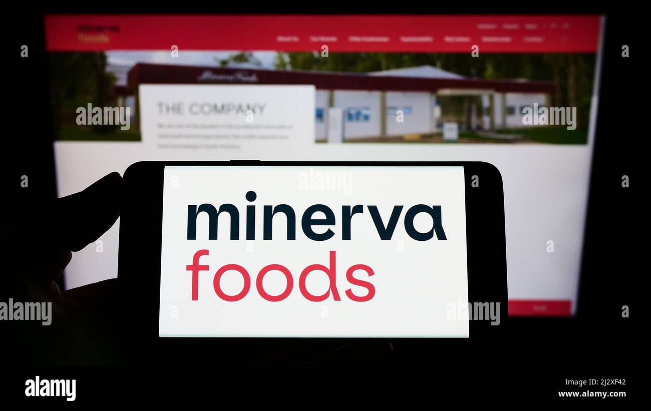 Person holding smartphone with logo of Brazilian foods company Minerva S.A. on screen in front of website. Focus on phone display. Stock Photo