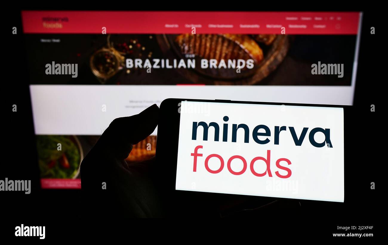 Person holding cellphone with logo of Brazilian foods company Minerva S.A. on screen in front of business webpage. Focus on phone display. Stock Photo