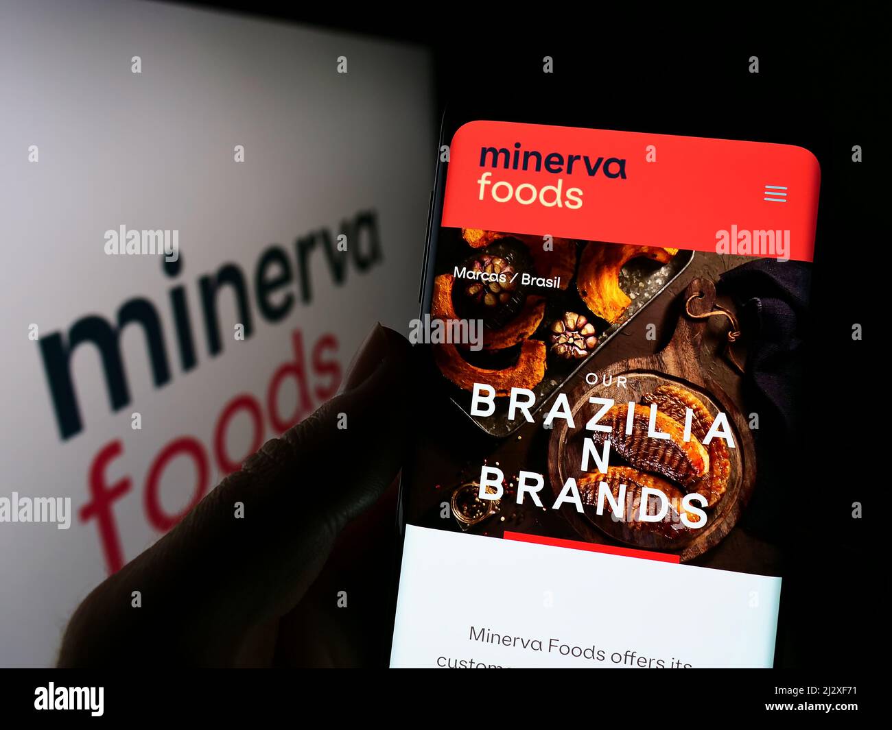 Person holding mobile phone with webpage of Brazilian foods company Minerva SA on screen in front of logo. Focus on center of phone display. Stock Photo