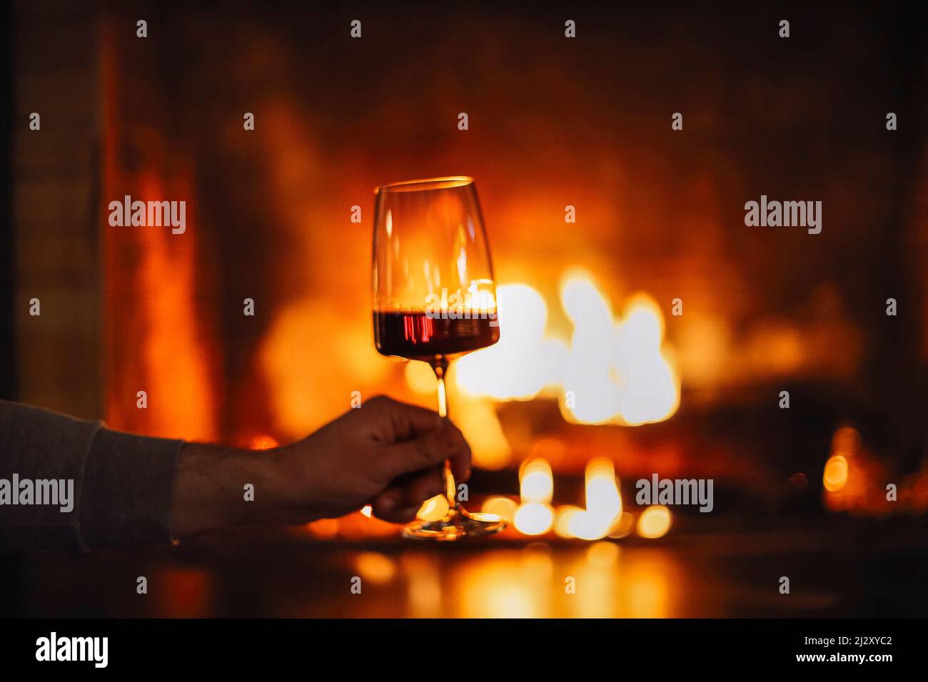 Man having red wine by the fireplace. Close up of man holding and drinking red wine glass by open wood fire place relaxing and enjoying quiet moment a Stock Photo