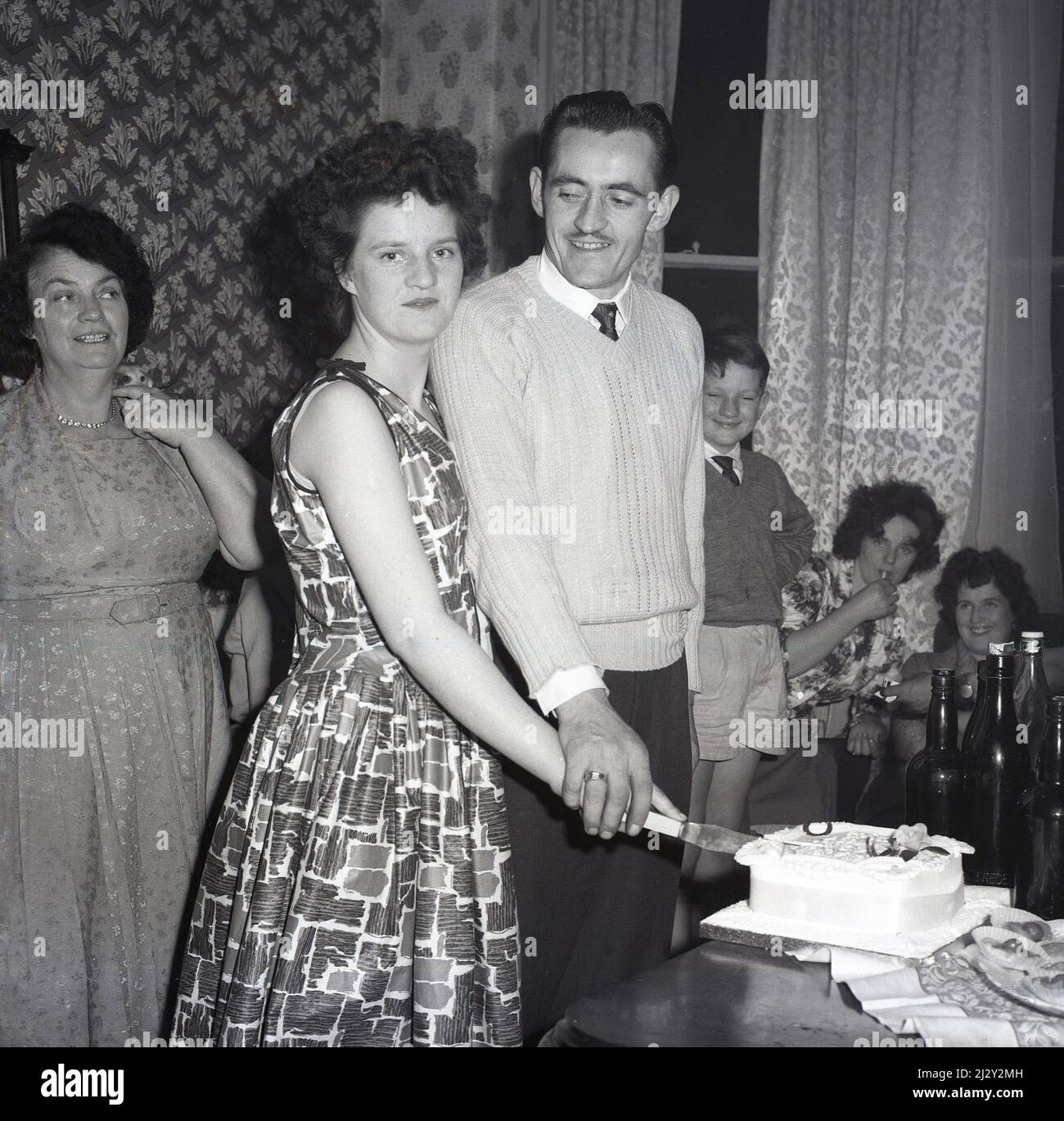1961, historical, engagement party, inside a room at a house, a young woman wearing a sleeveless dress of the era, with her older fiance, standing cutting their celebration cake together, Stockport, Manchester, England, UK. Stock Photo