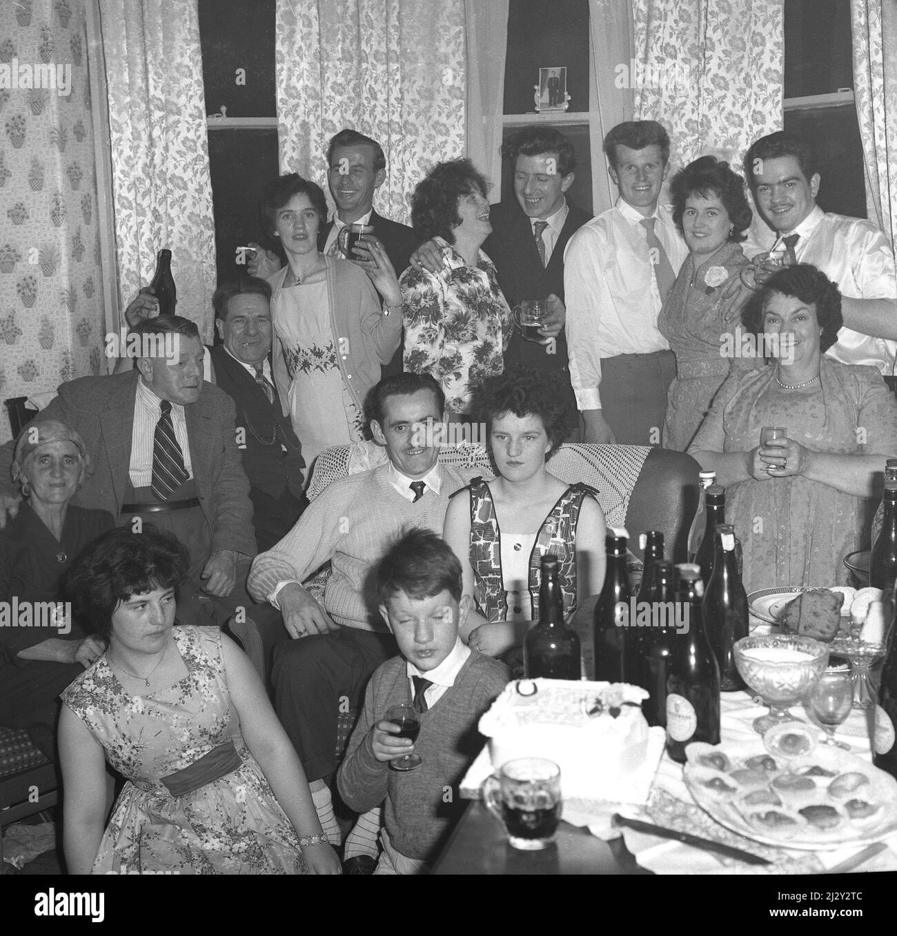 1961, historical, engagement party, engaged couple stting on a sofa chair together, surrounded by friends and family, Stockport, Manchester, England, UK. A celebratory cake is on the table as are bottles of beer, while sitting beside the table, sister gives her younger brother a look! Stock Photo