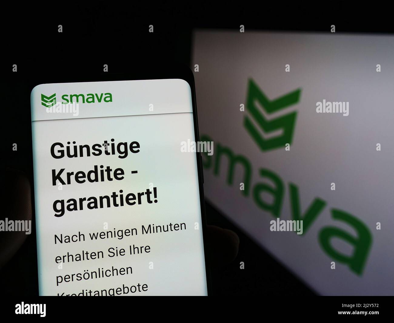 Person holding smartphone with website of German loan comparison company smava GmbH on screen in front of logo. Focus on center of phone display. Stock Photo