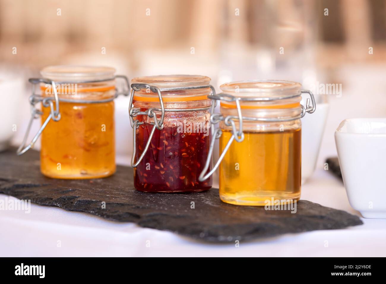 Three pots of conserves for toast or croissants in small glass jars on a breakfast table. The jars contain honey, marmalade and strawberry jam. Stock Photo