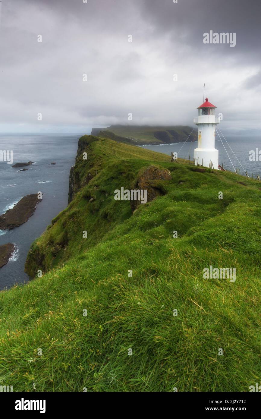 View of cliffs and Mykines Holmur Lighthouse up to Mykines, Faeroe Islands. Dark clouds. Stock Photo