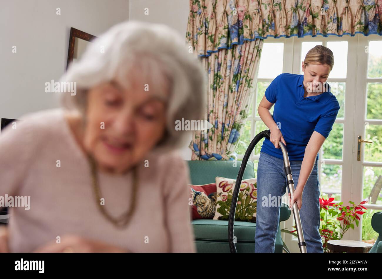 Female Home Help Cleaning House With Vacuum Cleaner And Talking To Senior Woman Stock Photo