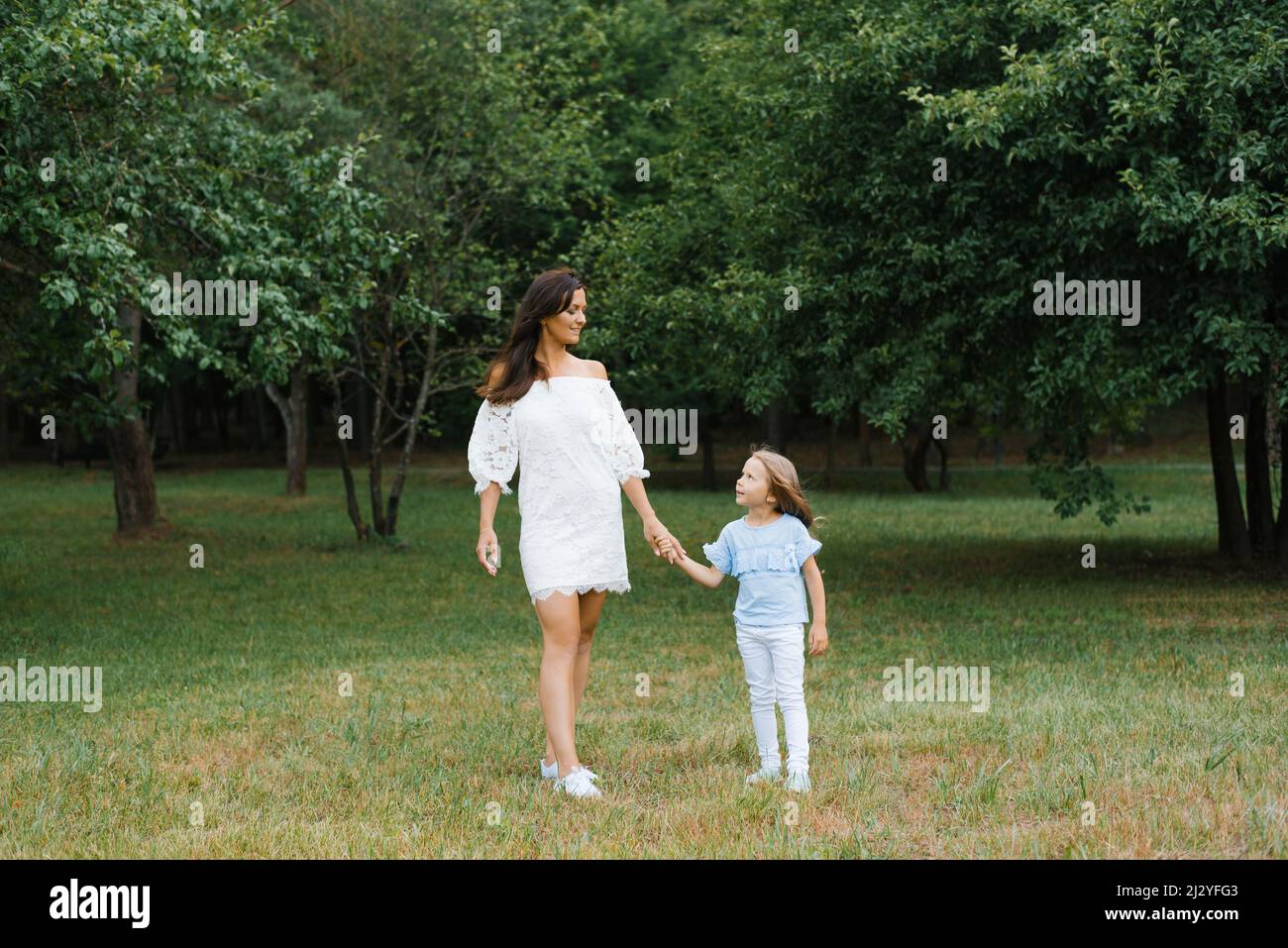 A young mother in a white dress holds her daughter's hand while walking in the park Stock Photo