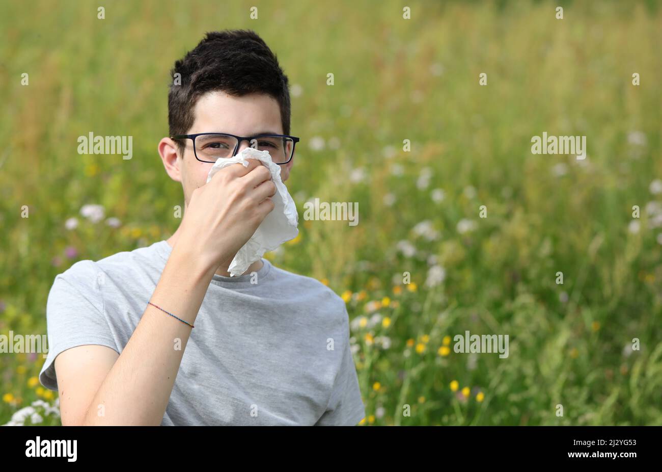 allergic boy with glasses sneezing on a white handkerchief outdoors because he has pollen allergy Stock Photo