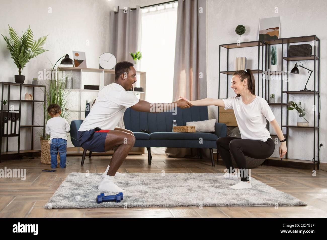 Young multinational multiracial couple doing squats holding hands training together at home, african man and caucasian woman working out together while their son is playing in the background. Stock Photo