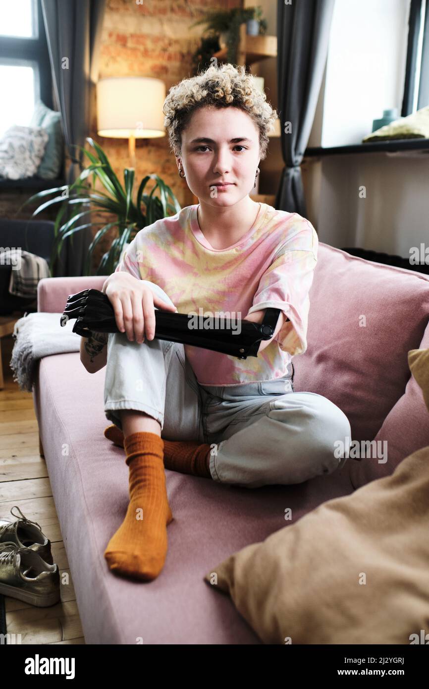 Portrait of girl with prosthetic arm in casual clothing sitting on sofa in living room and looking at camera Stock Photo