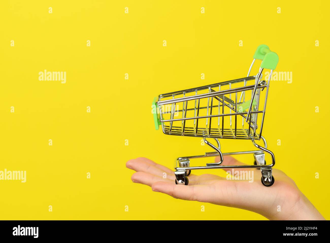 Empty shopping cart on female hand. Shopping theme. Small Trolley isolated on a bright yellow background. Consumer concept, Copy space. Stock Photo
