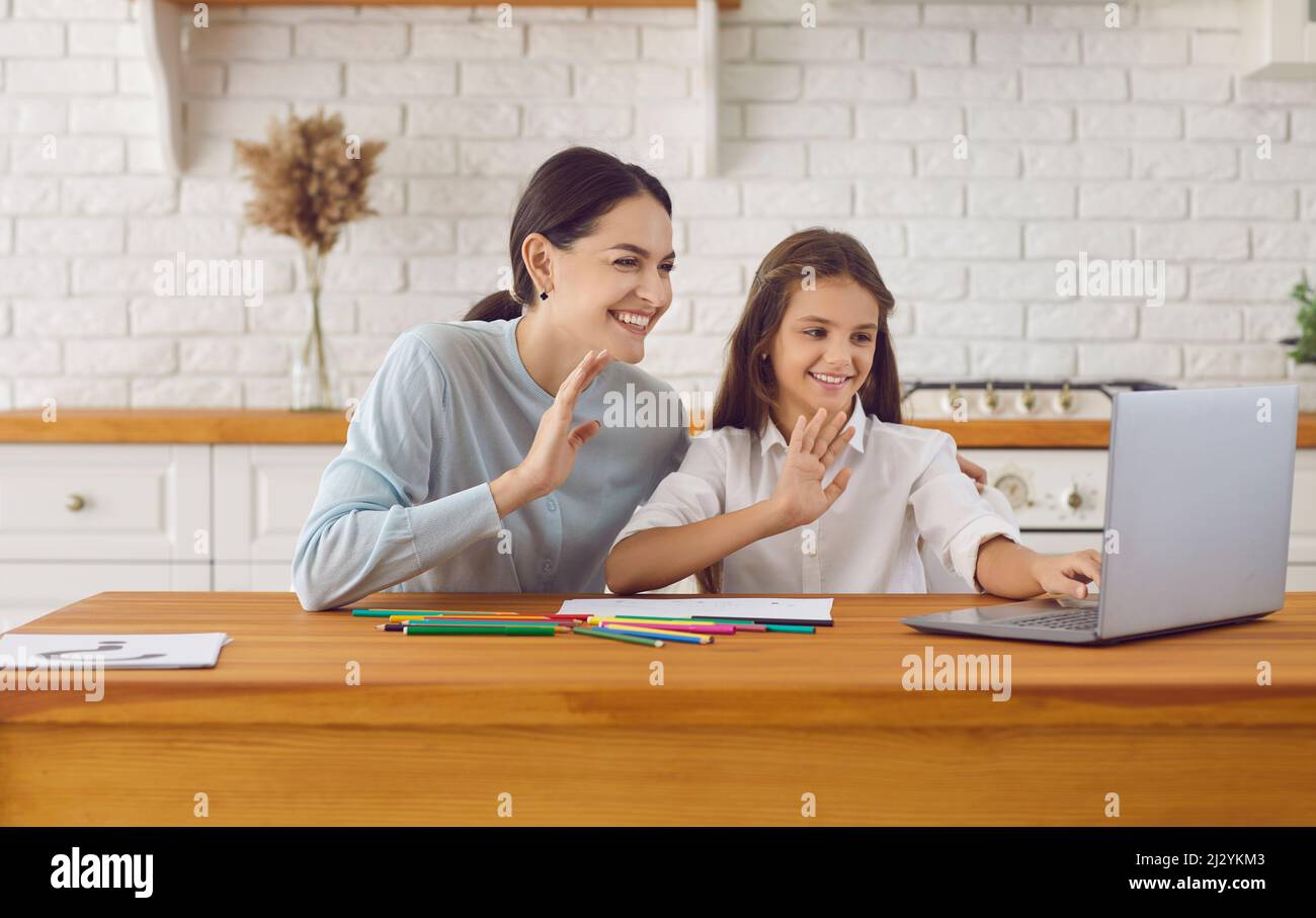 Happy mom and kid sitting at kitchen table and waving hello to teacher during virtual class Stock Photo