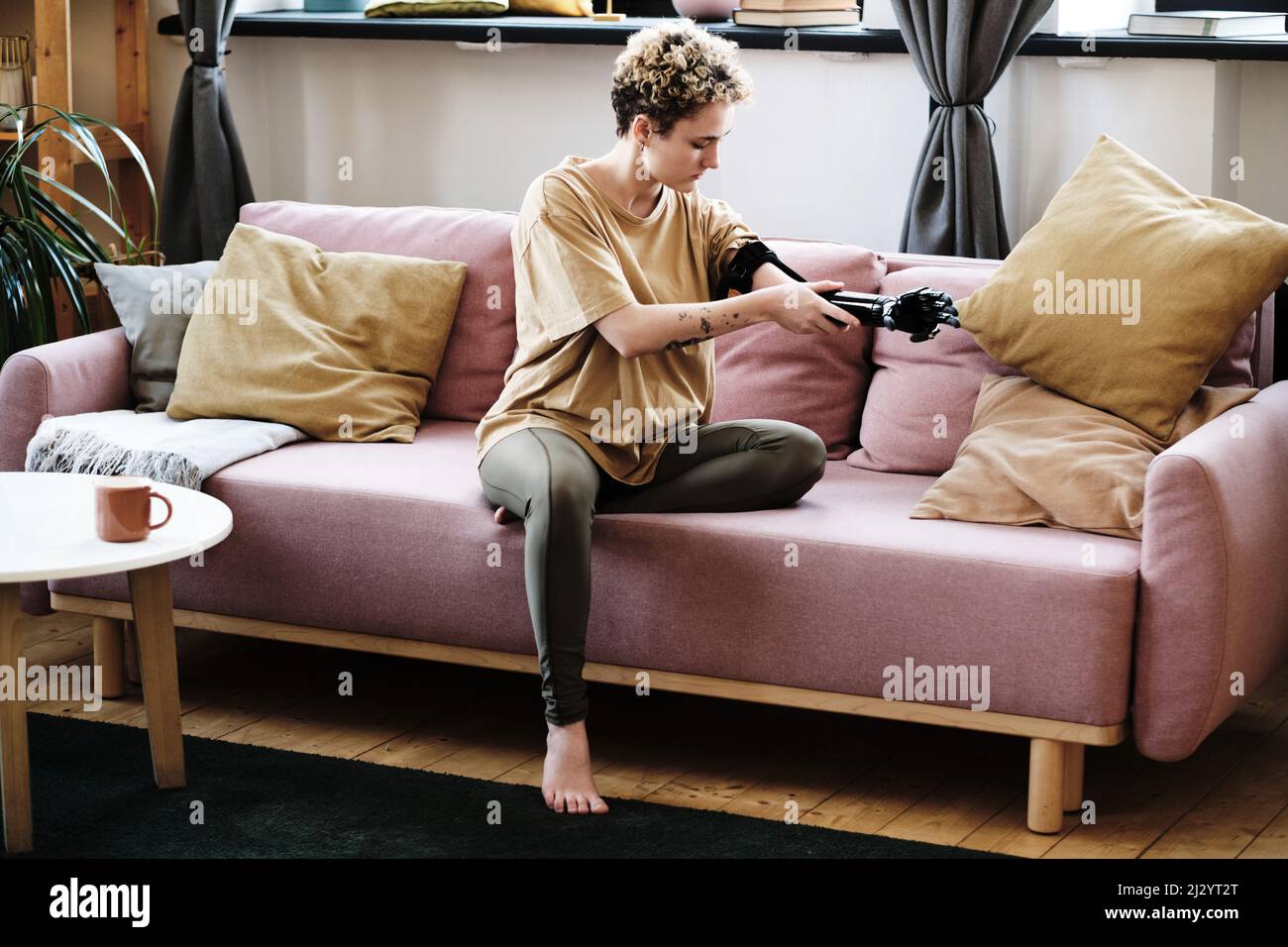 Young woman sitting on sofa in living room and putting on prosthetic arm Stock Photo