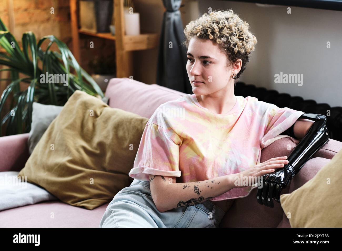 Young woman in casual clothing with prosthetic arm resting on sofa in living room Stock Photo