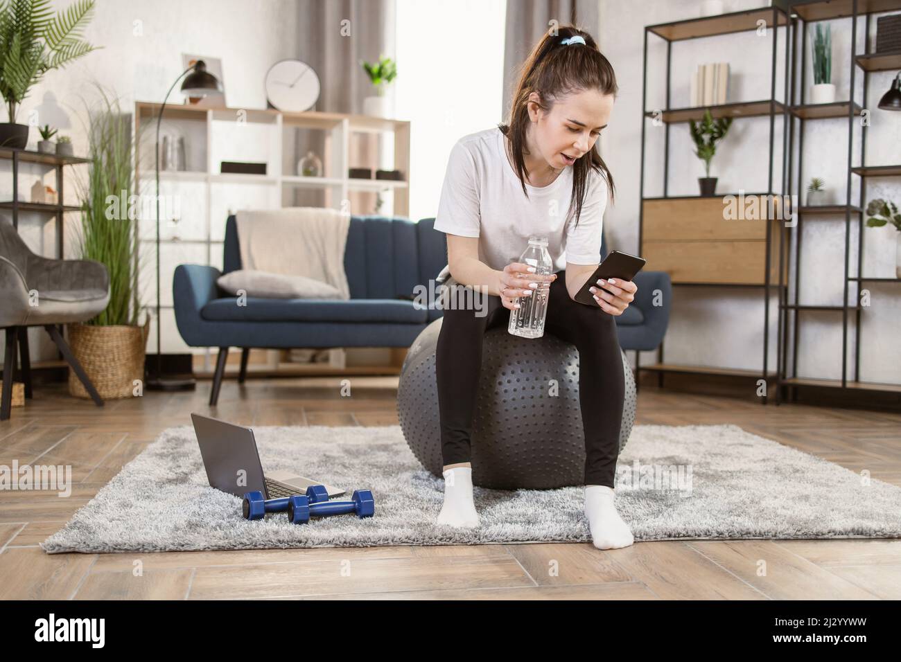 Happy fitness woman training on swiss ball in living room. Portrait of muscular woman enjoying home fitness activity , sitting on rubber fitball, smiling, drinking water and looking at his cell phone Stock Photo