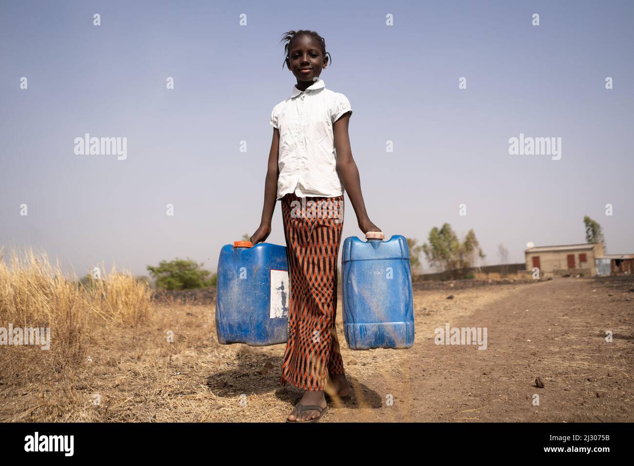 Skinny African girl carrying two water containers in the middle of an arid landscape; concept of poverty, lack of pipe connections in private rural ho Stock Photo