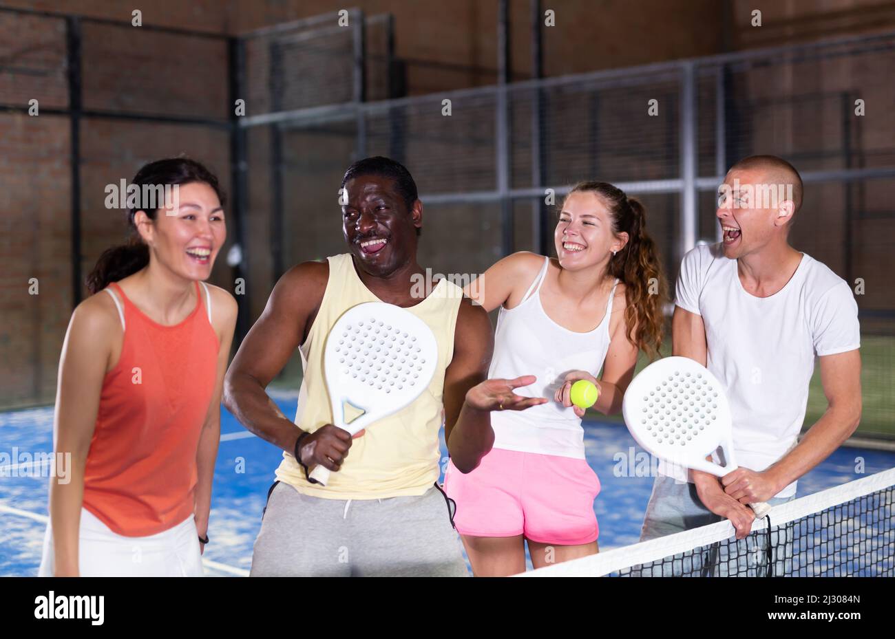 Laughing men and women with rackets and balls talking on indoor padel court Stock Photo