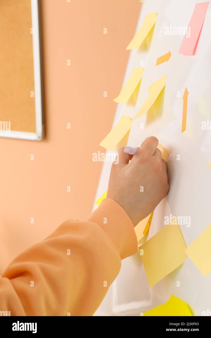 Woman writing on sticky note attached to flipchart near beige wall, closeup Stock Photo