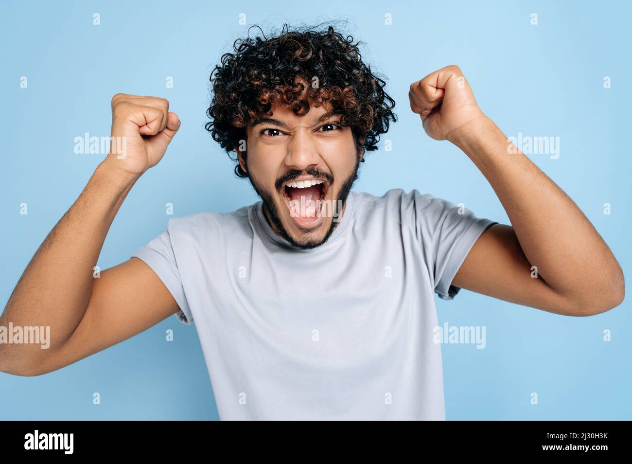 Excited amazed indian or arabic guy, in a basic t-shirt, rejoices in success, victory, win, gesturing with fists, looking at the camera, smiling, standing on an isolated blue background, shouting Stock Photo