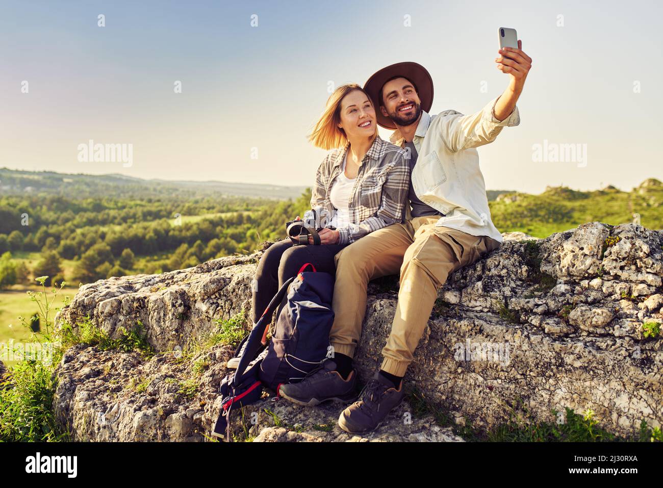 Young couple taking selfie while sitting on rock in mountains during summer hike Stock Photo