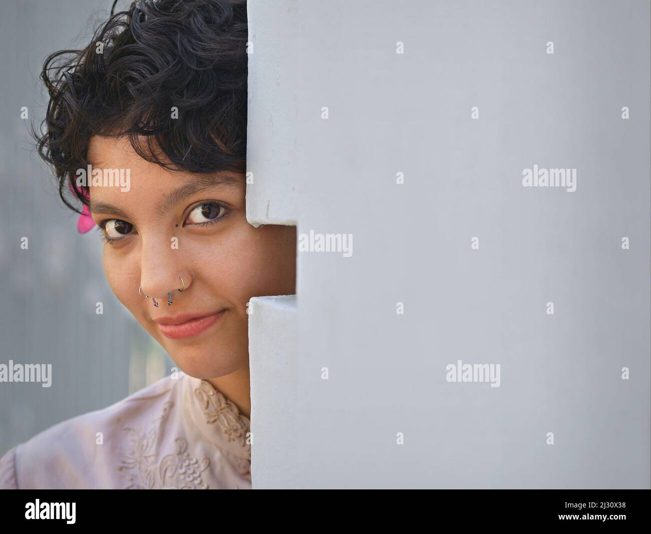 Shy pretty young Latina woman with beautiful big brown eyes and short curly hair looks cautiously around a corner towards the viewer. Stock Photo