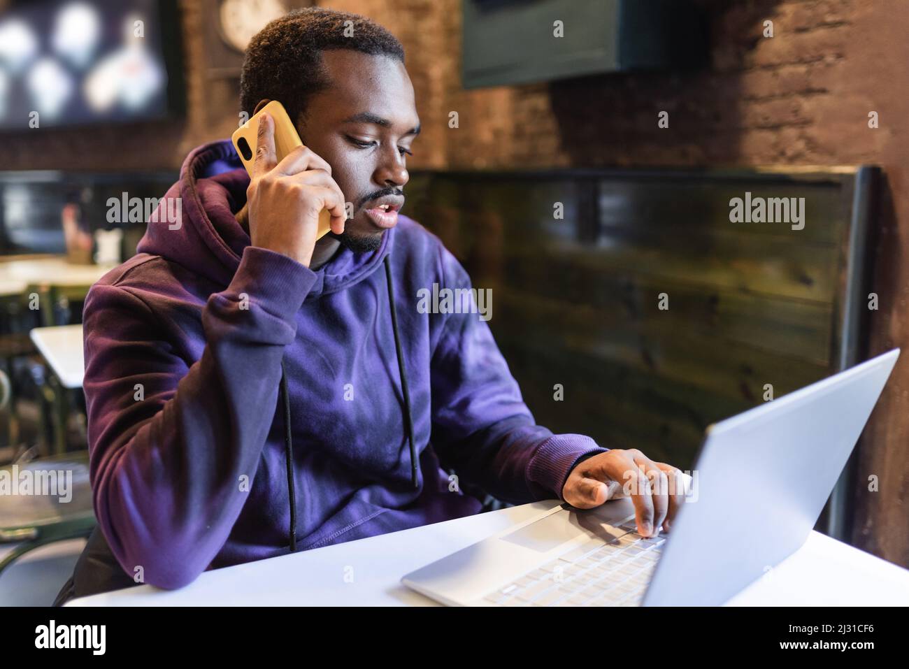 Young African American male freelancer talking on cellphone while doing remote job on netbook in cafe Stock Photo