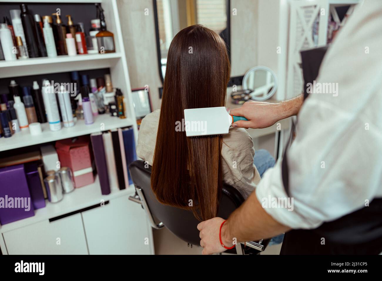 Hairstylist brushing long and sleek brown hair of female client at beauty salon Stock Photo