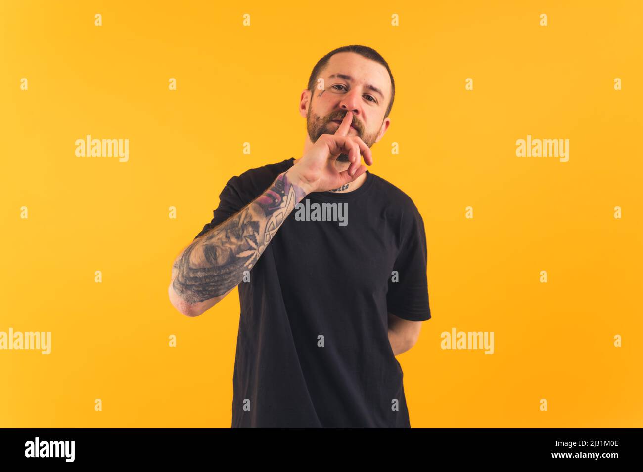 young Caucasian man wearing a black T-shirt and with a big tatto on his hand putting a finger on mouth and looking into the camera yellow background studio shot medium shot . High quality photo Stock Photo