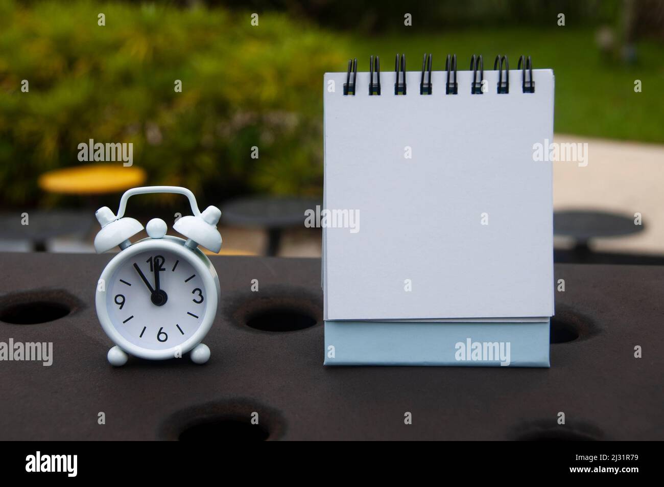 Alarm clock pointing at 11 o'clock with blank notepad. Blurred park background. Copy space Stock Photo