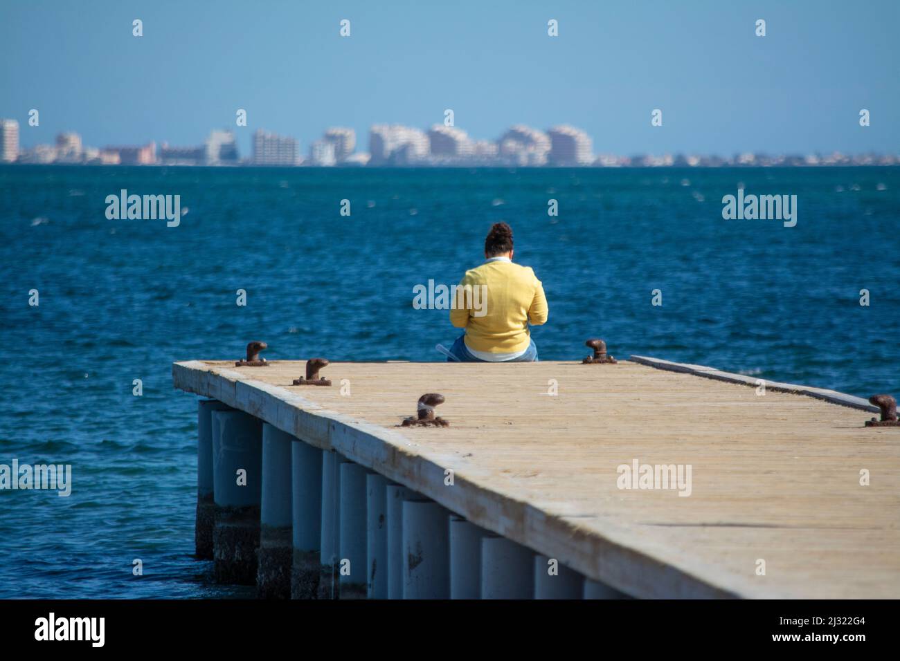 Person in a yellow shirt sitting on the edge of a jetty in the Mar Menor Stock Photo