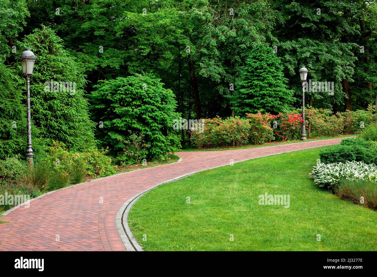 curved pedestrian pavement of stone tiles in park with slope landscape lighting and green plants on flower bed, meadow of on turf lawn with pillar gar Stock Photo