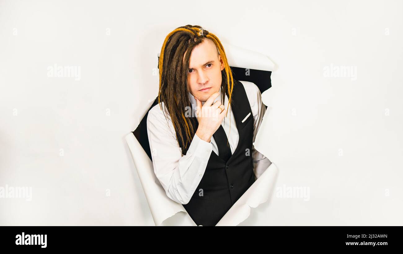 Young man with dreadlocks scratching chin. Handsome male in white shirt, black vest and tie with thoughtful look in hole of white background Stock Photo