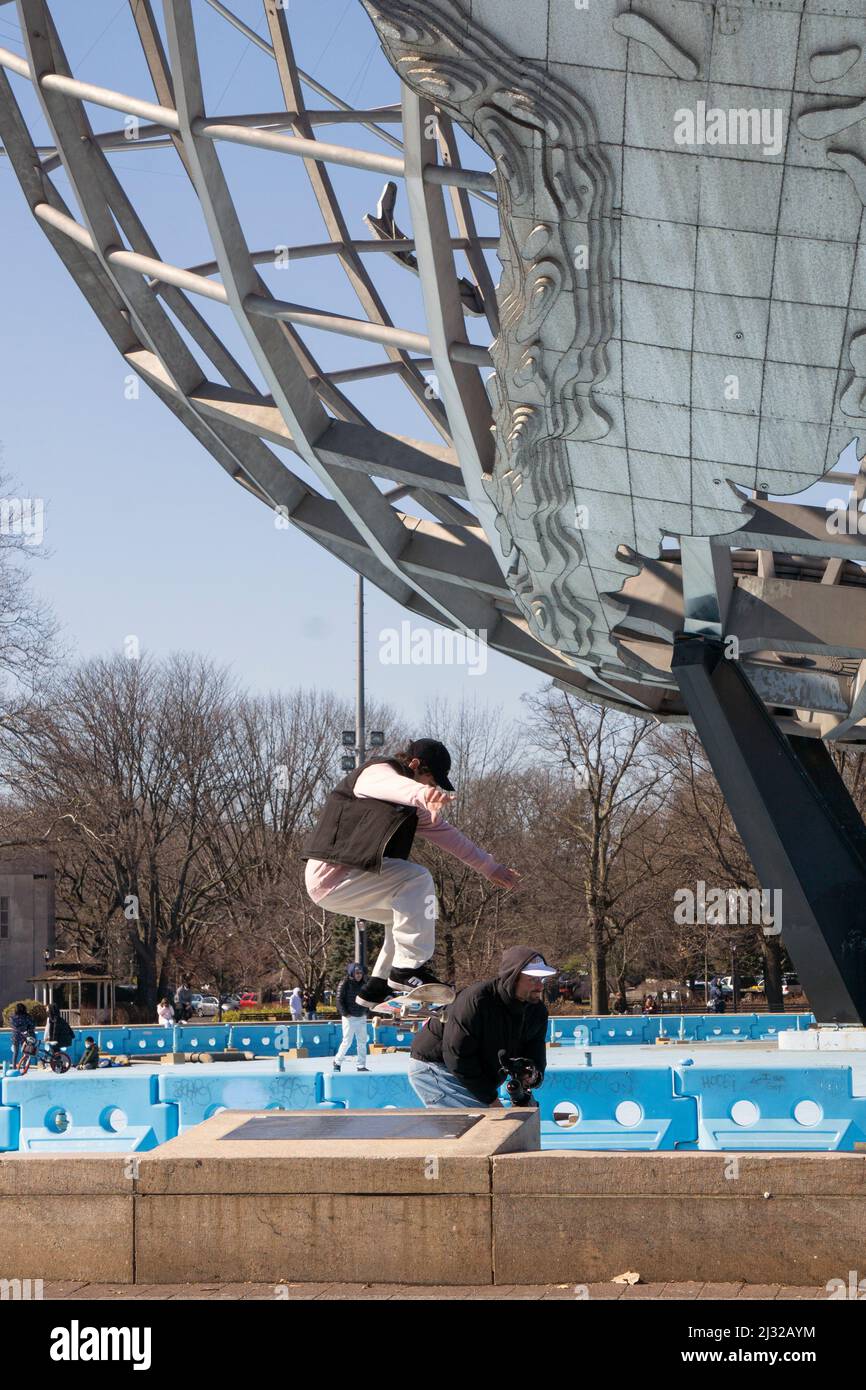 In the shadow of the Unisphere, a stunt skater jumps over an obstacle while a fellow skater films him. In a park in Queens, New York City. Stock Photo