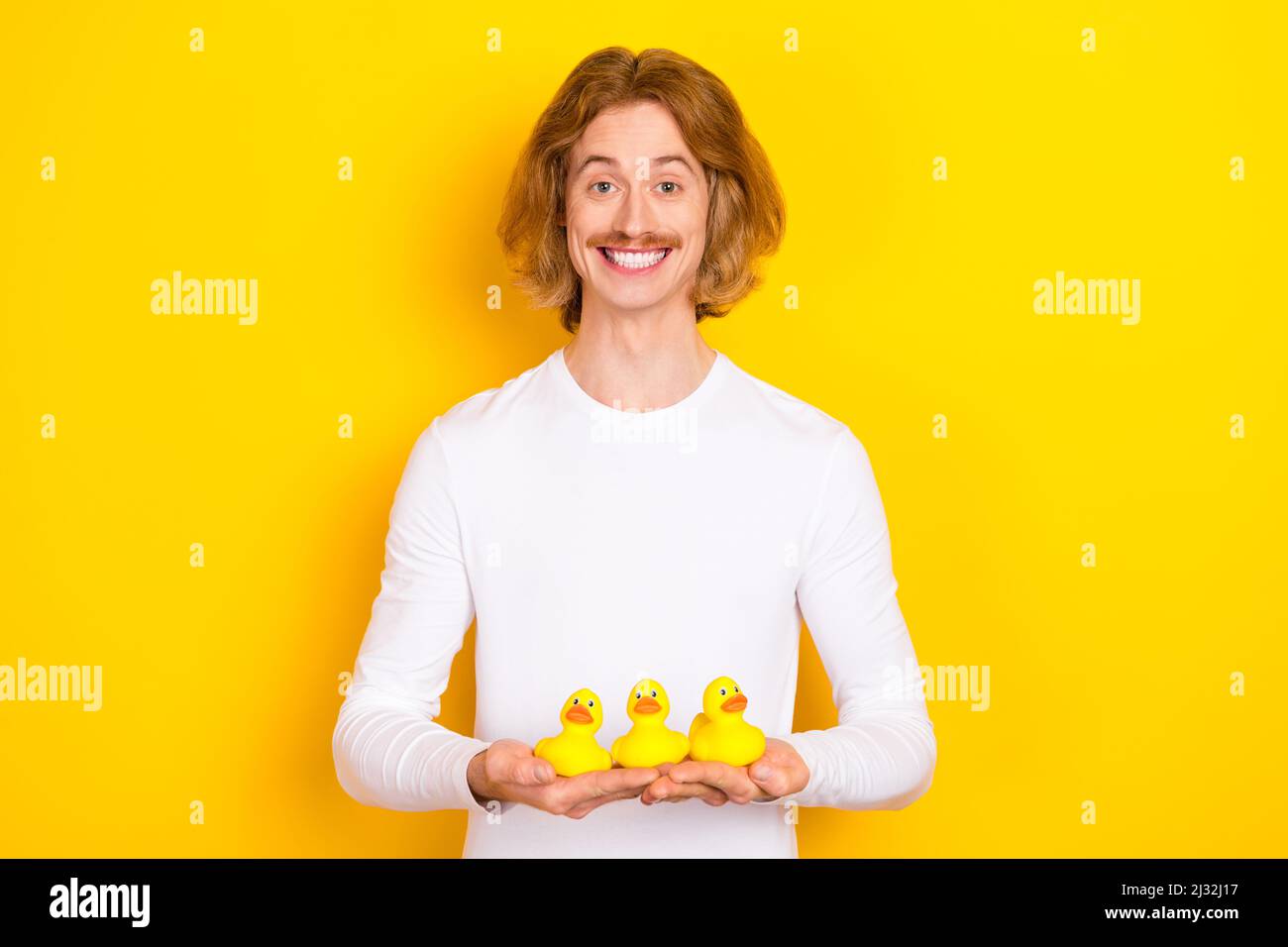 Portrait of handsome cheerful funny childish male take bath holding three little ducklings isolated on yellow color background Stock Photo